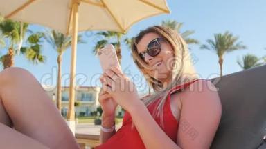 穿红色比基尼的女孩在度假时使用她的智能手机在酒店泳池边区域。 度假旅行妇女在水疗中心放松